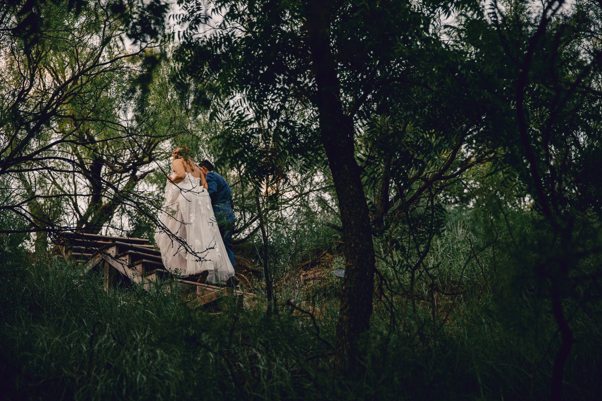 A bride and groom are kissing on a bridge in the woods.
