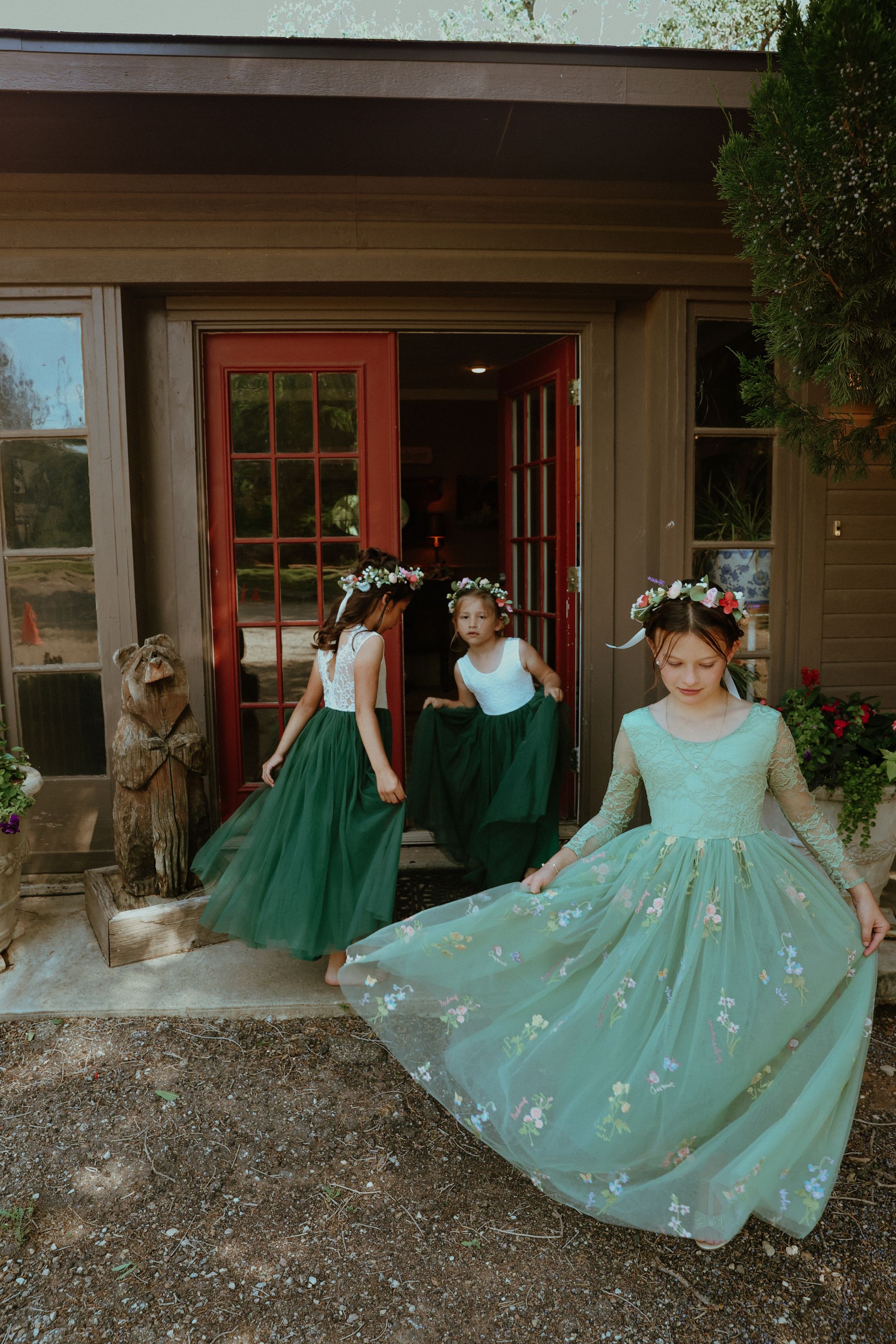 Three flower girls in green dresses are standing in front of a house.