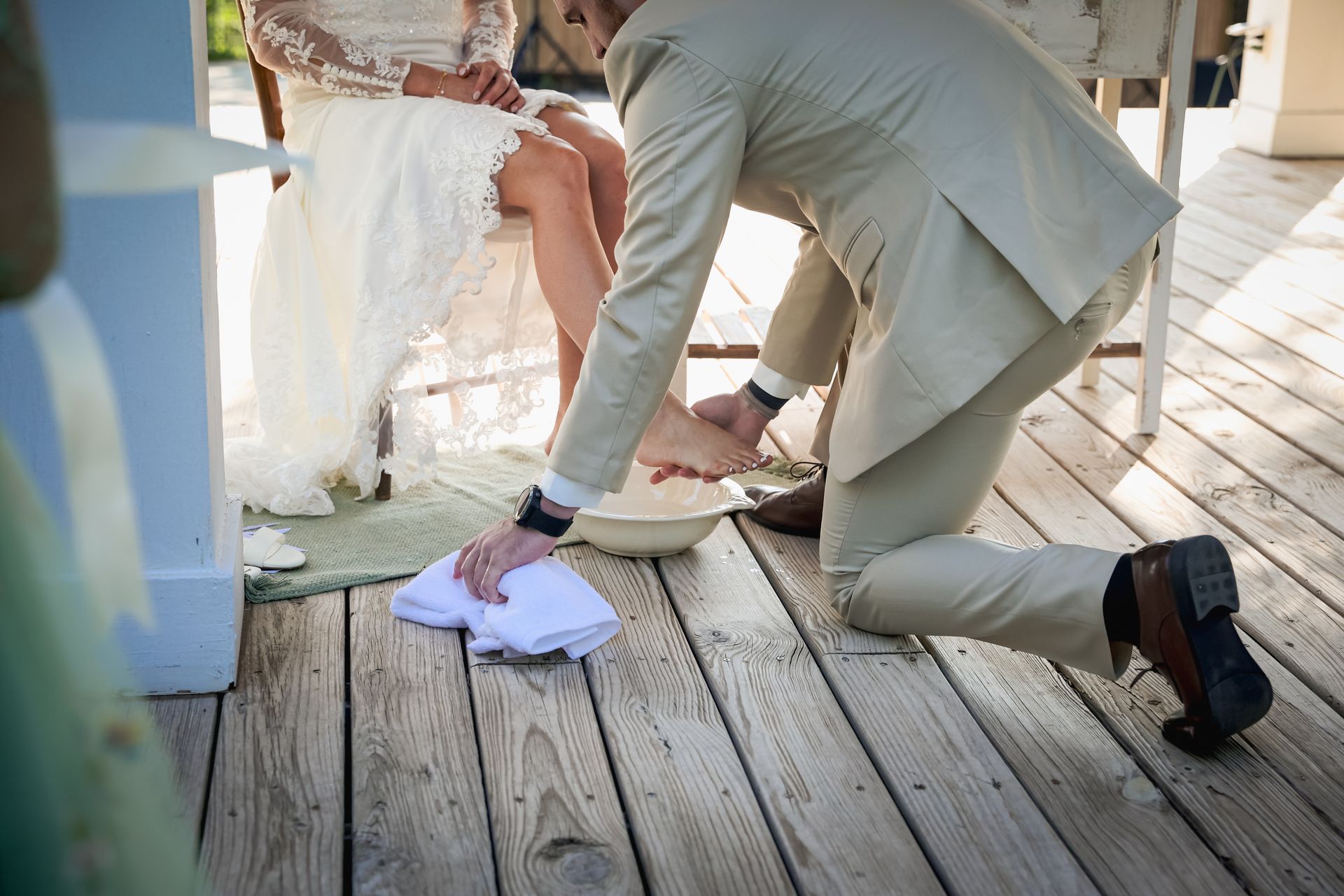A bride and groom are sitting on a wooden deck and the groom is washing the bride 's feet.