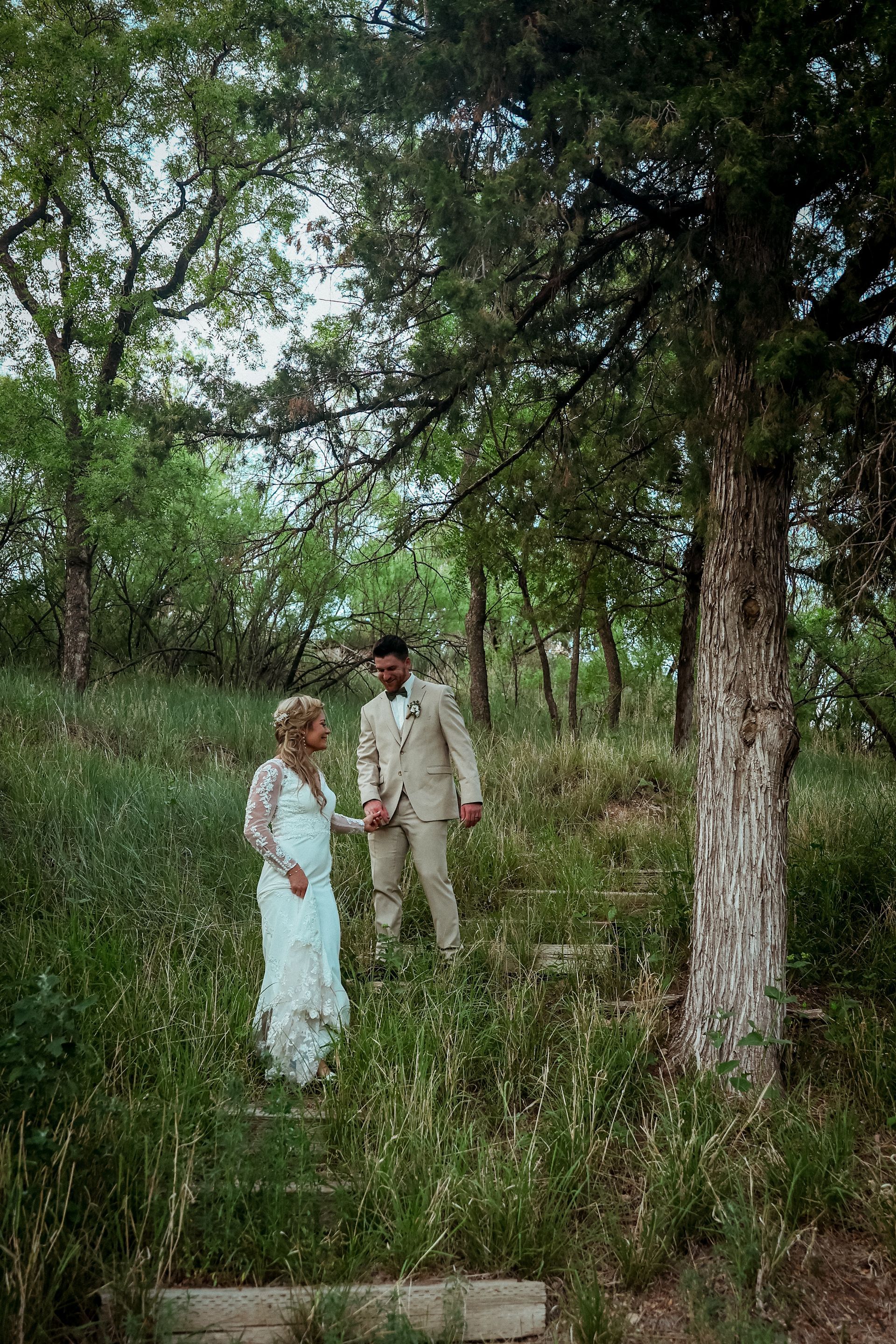 A bride and groom are walking through a forest holding hands.
