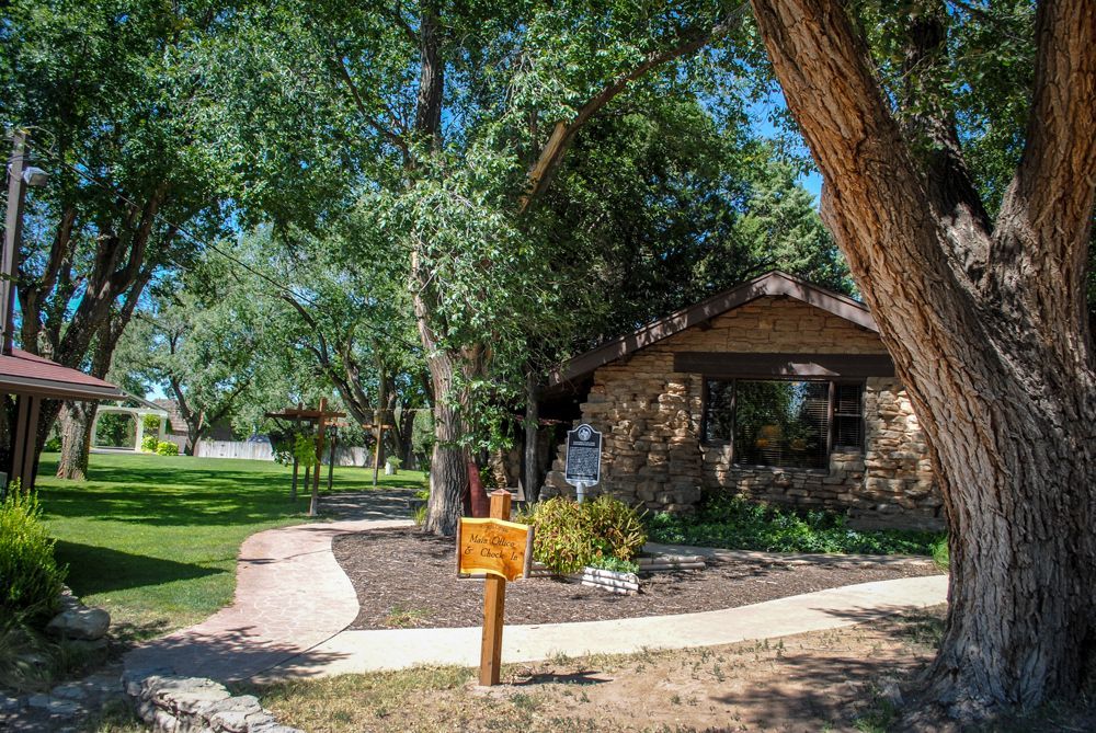 A wooden sign is in front of a stone house surrounded by trees.