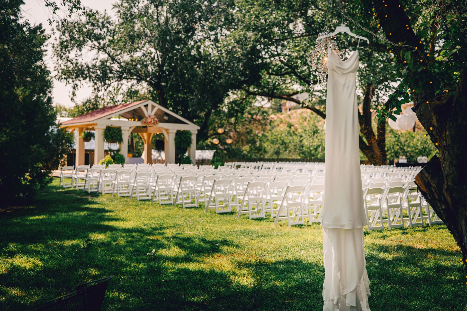 A wedding dress is hanging from a tree in front of a row of white chairs.