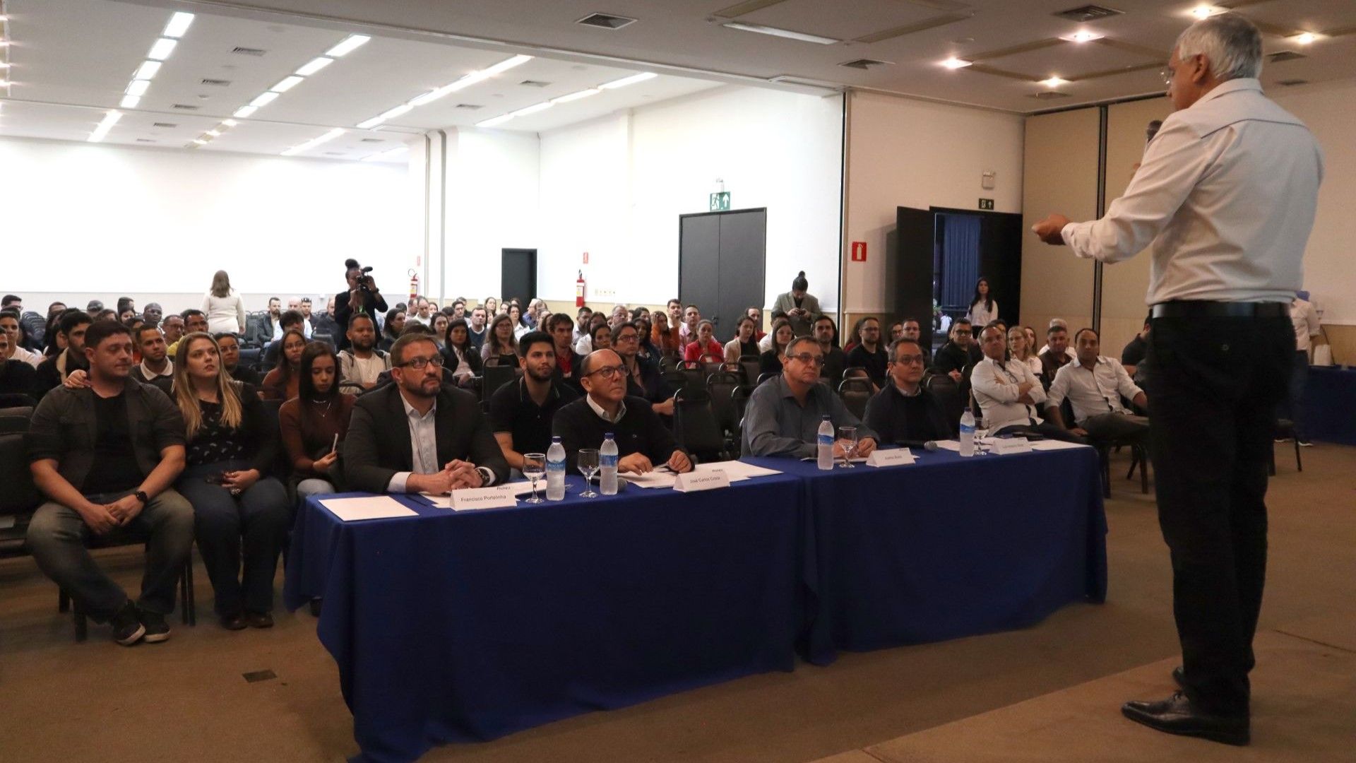 Um homem está de pé na frente de um grande grupo de pessoas sentadas em mesas.