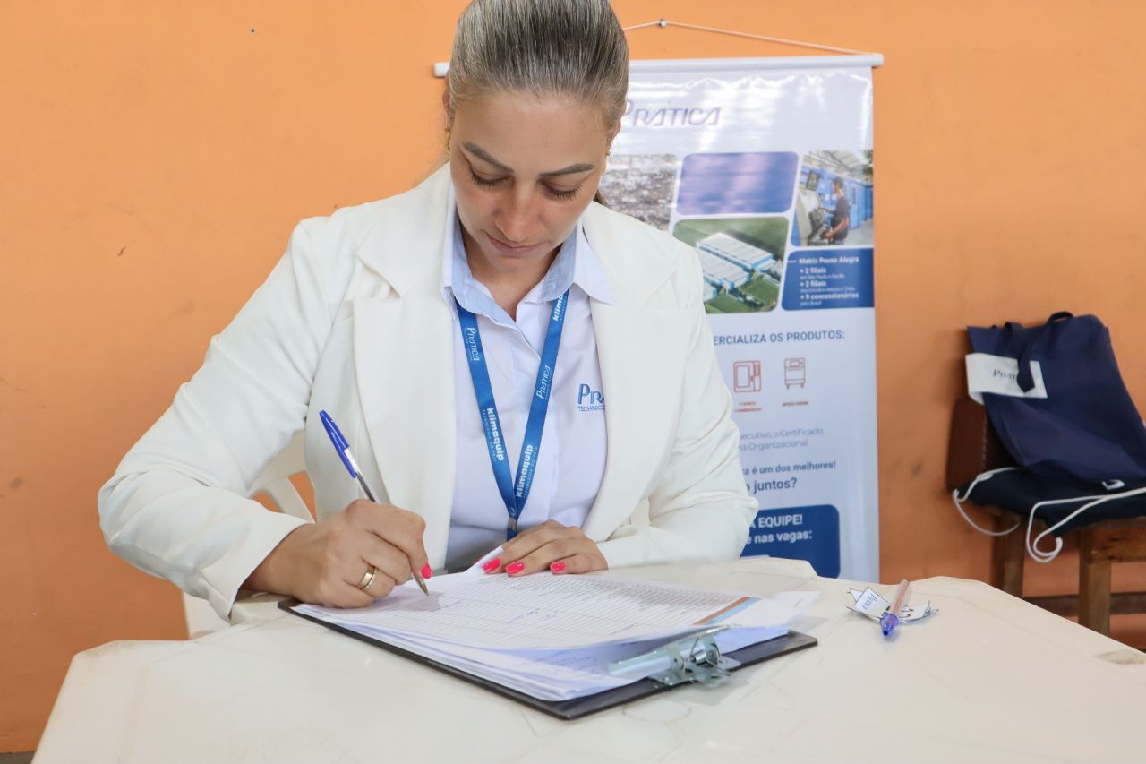 Uma mulher de casaco branco está sentada à mesa assinando um documento.