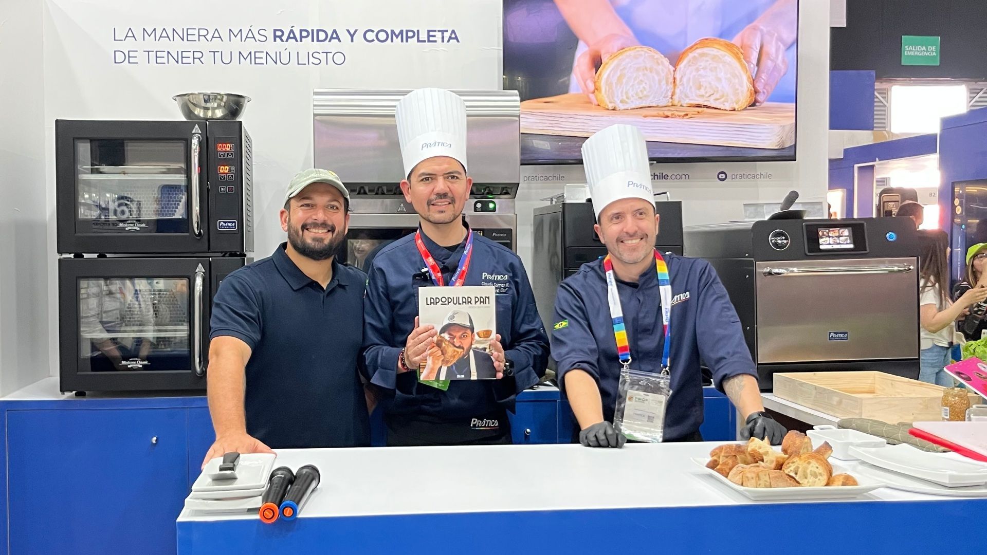 Três chefs em uma demonstração de culinária segurando um livro, com fornos atrás deles.