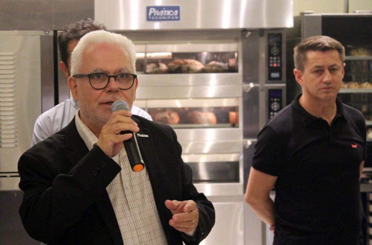Um homem de terno fala ao microfone em frente a um forno de aço inoxidável.
