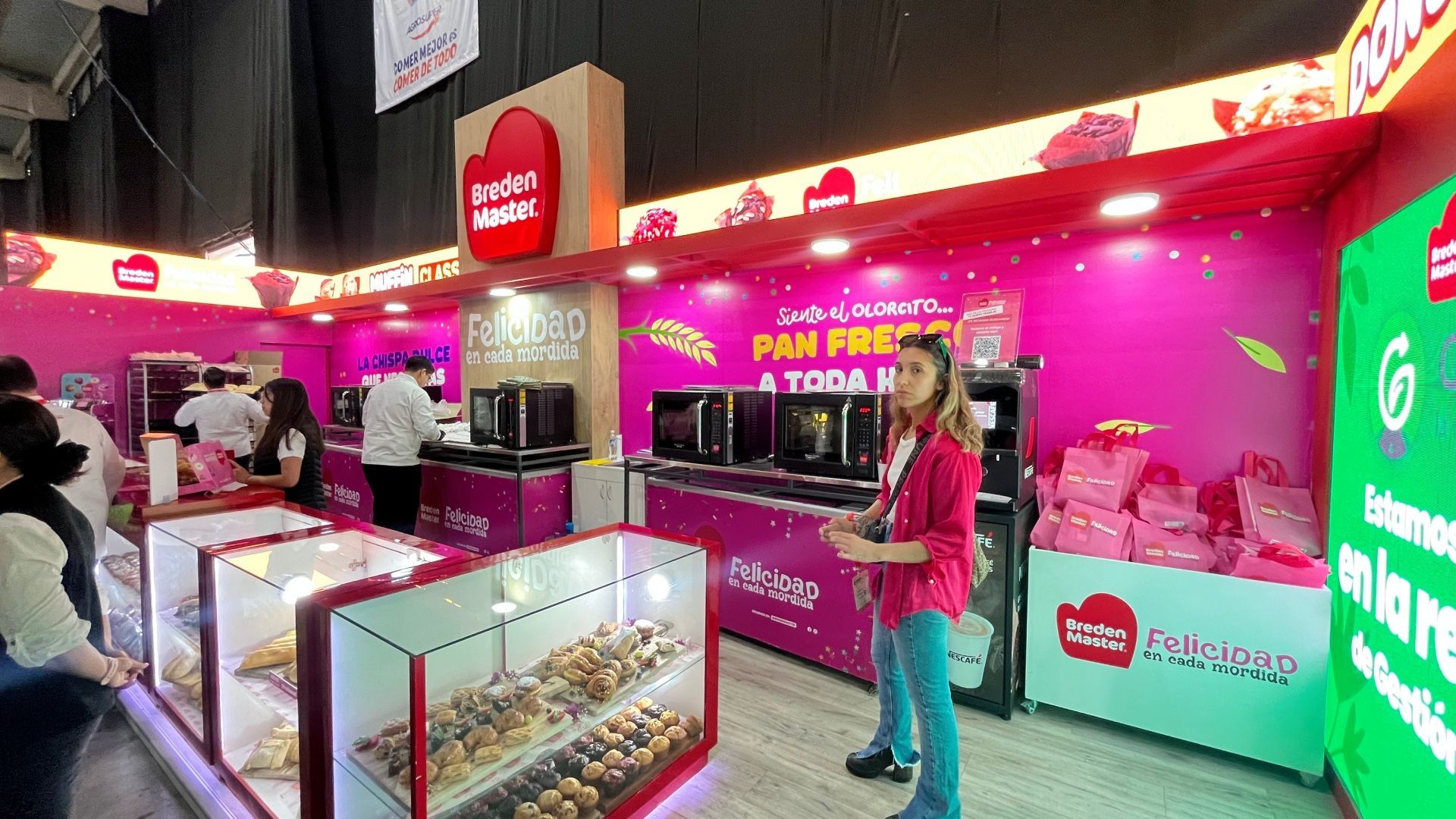 Barraca de padaria com detalhes em rosa, exibindo pães e doces. Uma mulher está perto da vitrine.