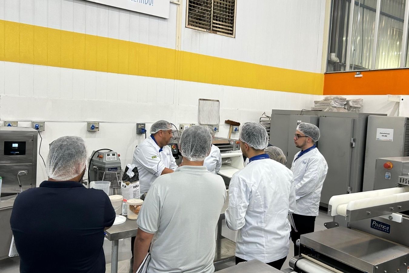 Grupo de pessoas com toucas e jalecos em uma área de produção de alimentos, ouvindo um homem.