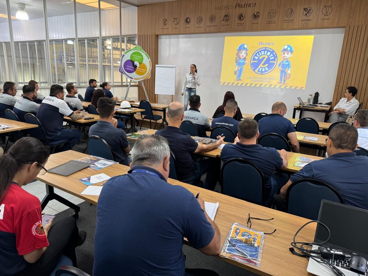 Um grupo de pessoas está sentado em mesas em uma sala de aula assistindo a uma apresentação.