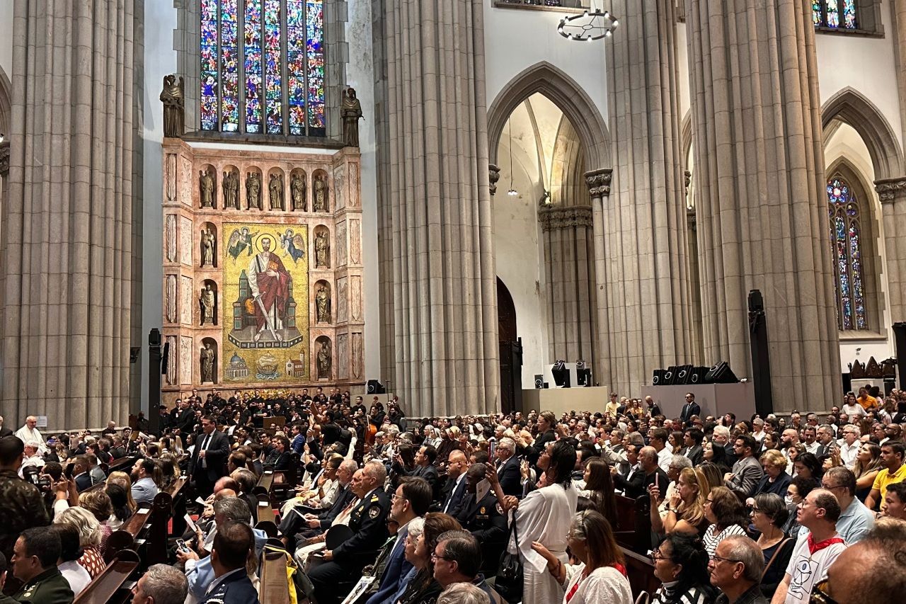 Grande multidão dentro de uma catedral, de frente para um altar com vitrais. Muitas pessoas estão sentadas.