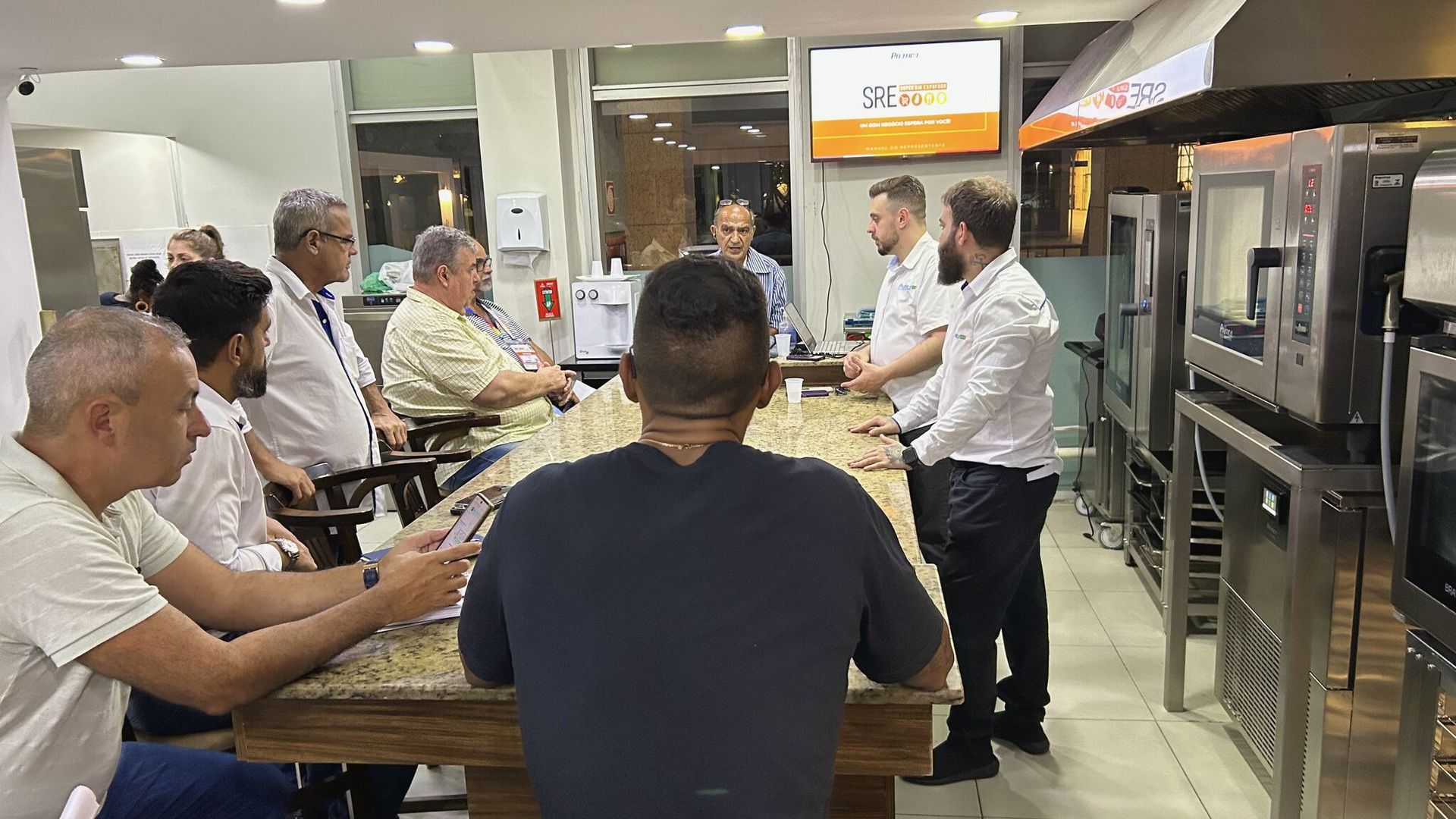 Um grupo de homens está em pé ao redor de uma mesa na cozinha.