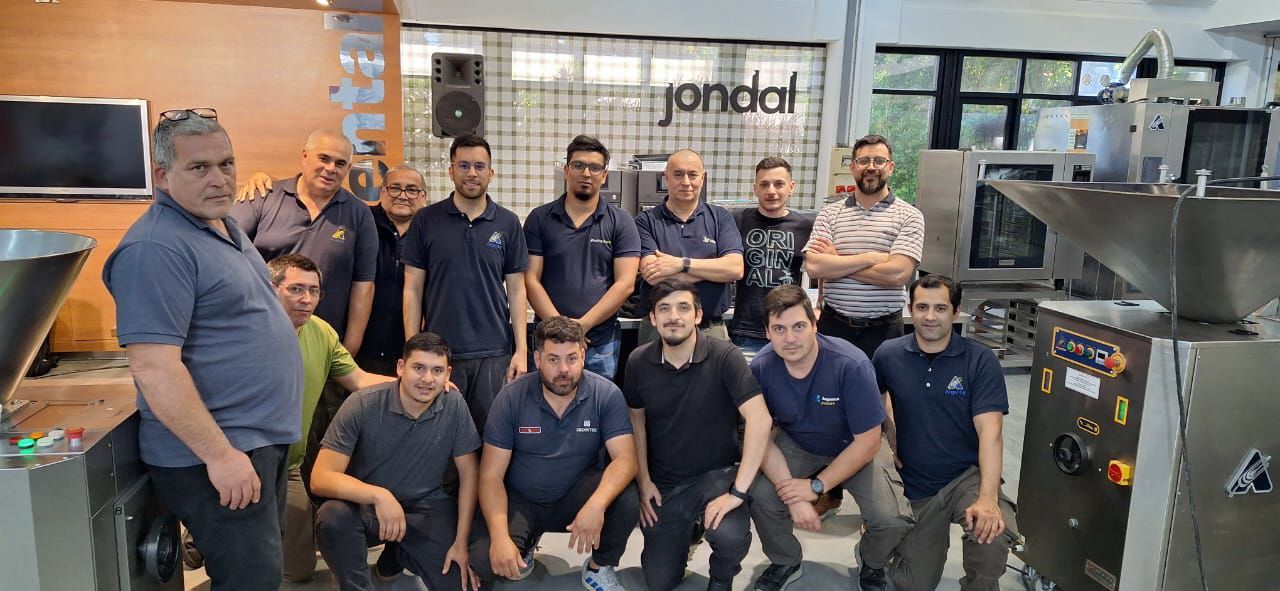 Um grupo de homens posando em uma oficina com máquinas. Alguns estão de pé, outros ajoelhados. O logotipo da empresa