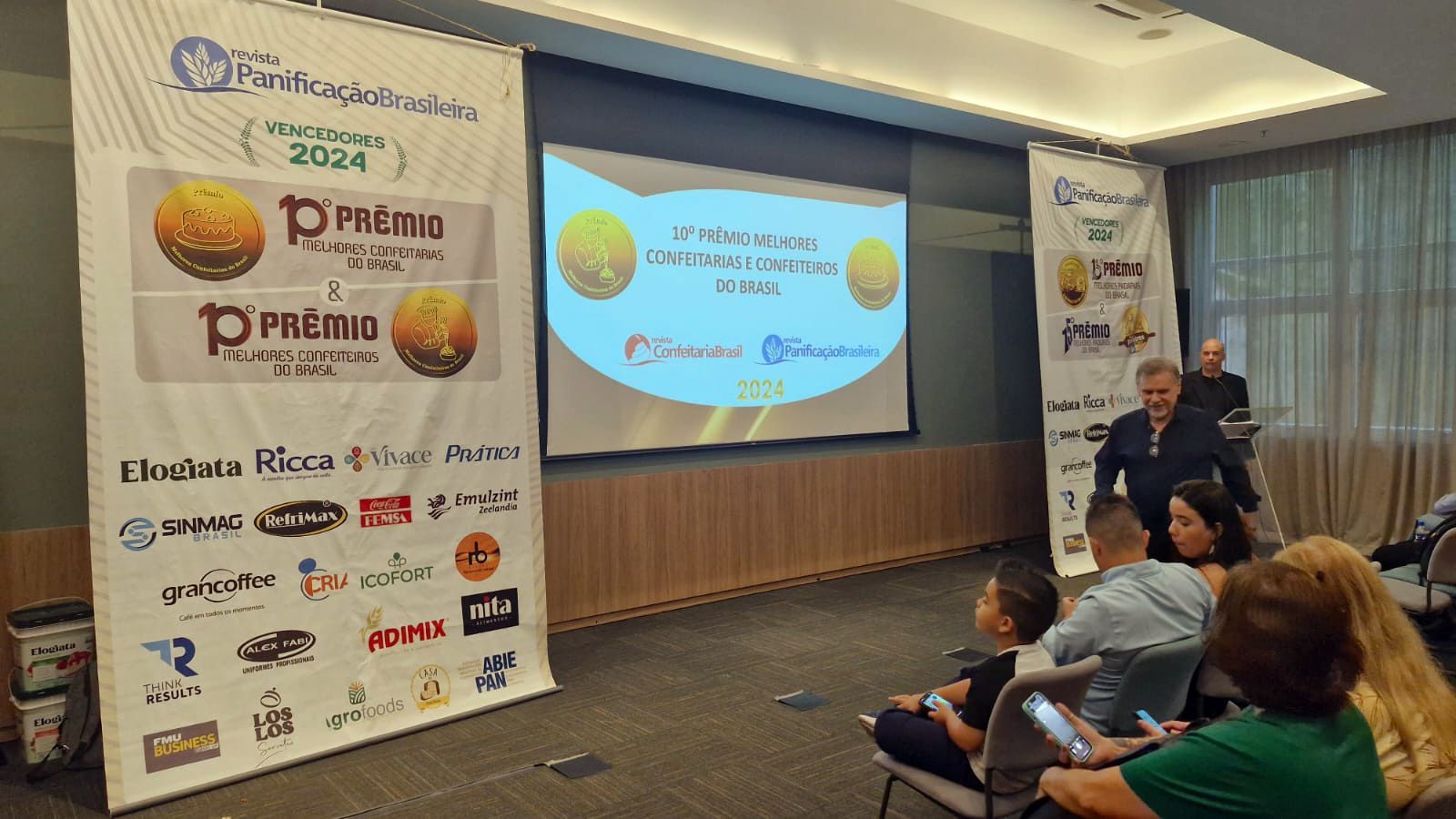 Um grupo de pessoas está sentado em frente a uma tela grande em uma sala.