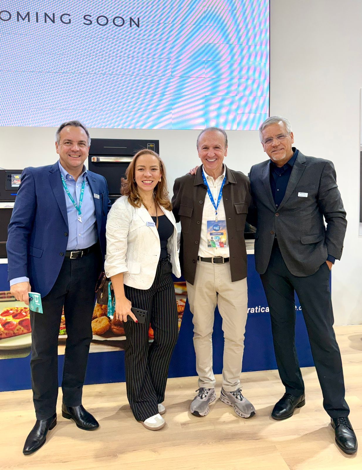 Quatro pessoas estão juntas em um ambiente fechado, perto de uma tela. Dois homens usam jaquetas, uma mulher usa um cardigã e a quarta pessoa usa uma jaqueta marrom.