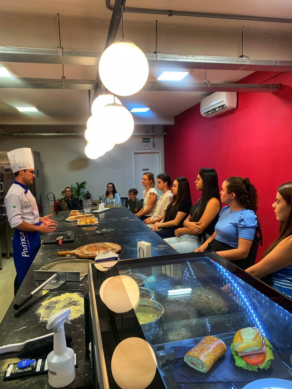 Um grupo de pessoas está sentado à mesa na cozinha enquanto um chef conversa com elas.