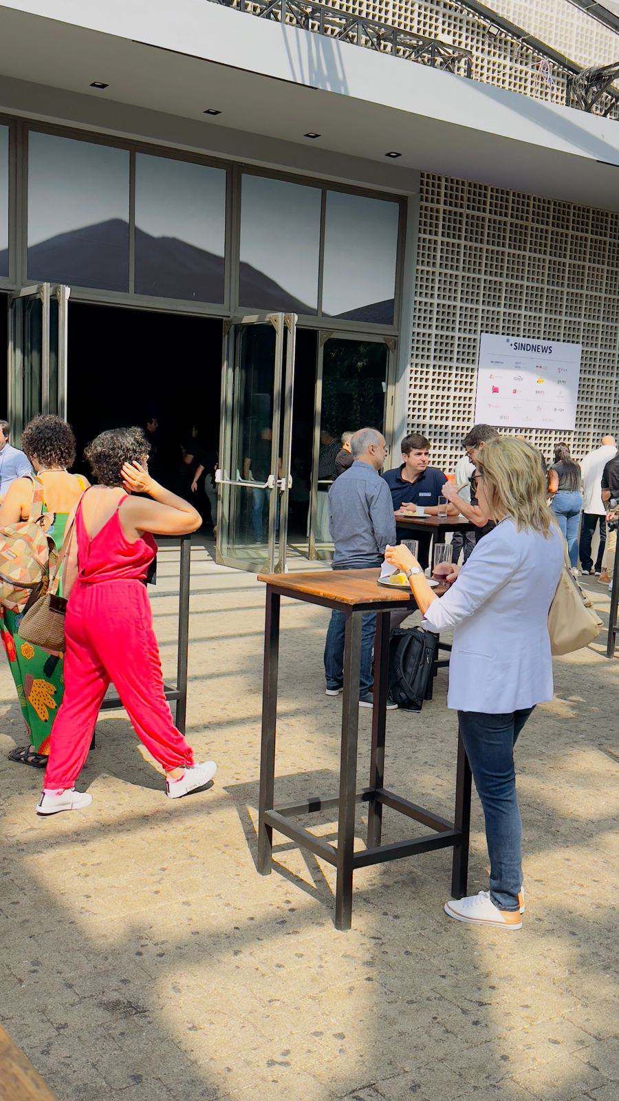 Um grupo de pessoas está de pé ao redor de mesas do lado de fora de um prédio.