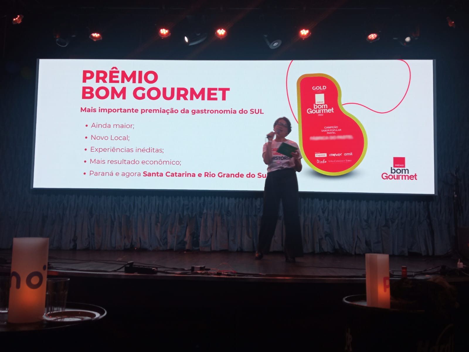 Uma mulher está parada em frente a uma tela grande que diz premio bom gourmet
