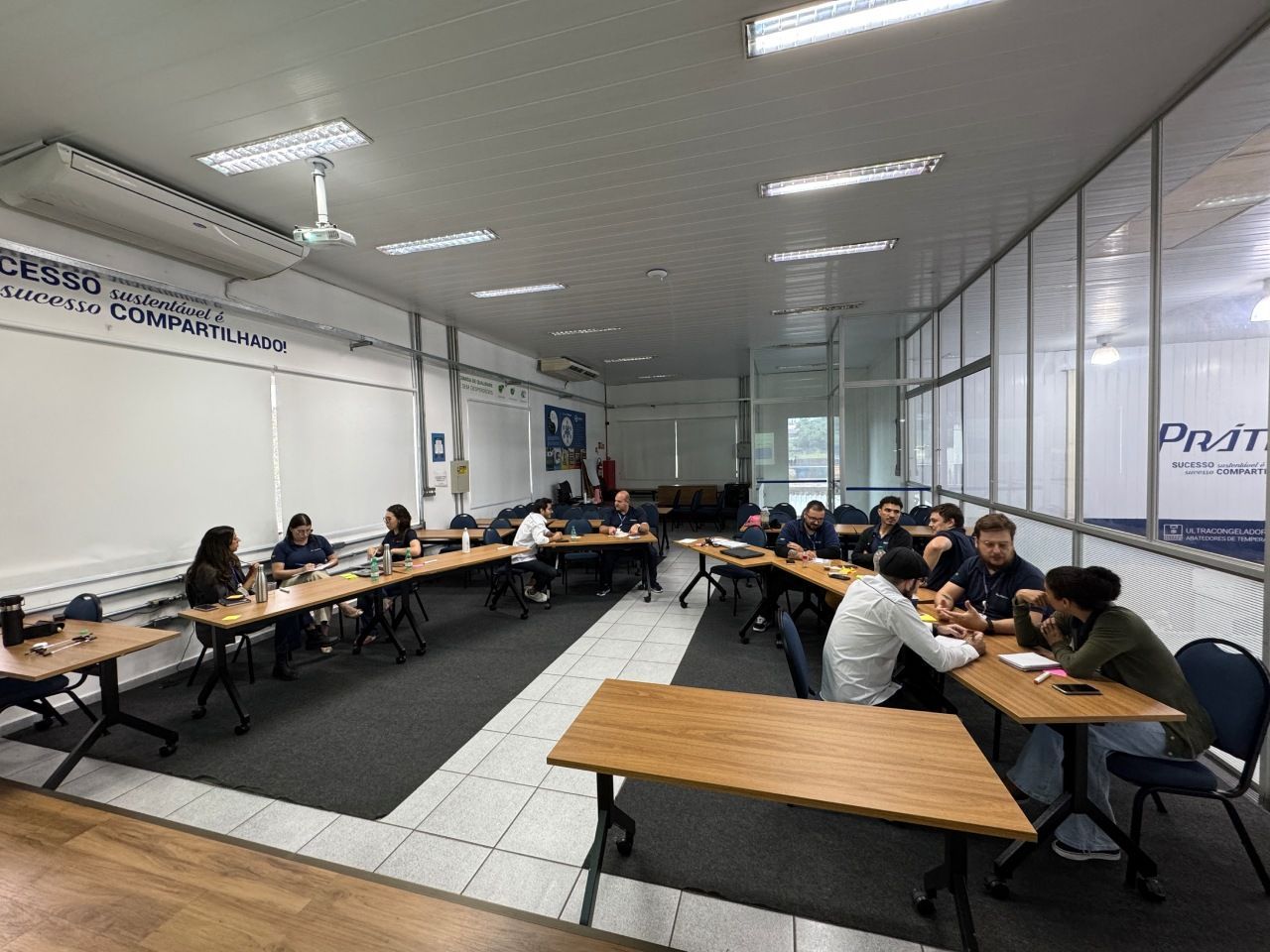Um grupo de pessoas está sentado em mesas dispostas em forma de U em uma sala de reuniões bem iluminada