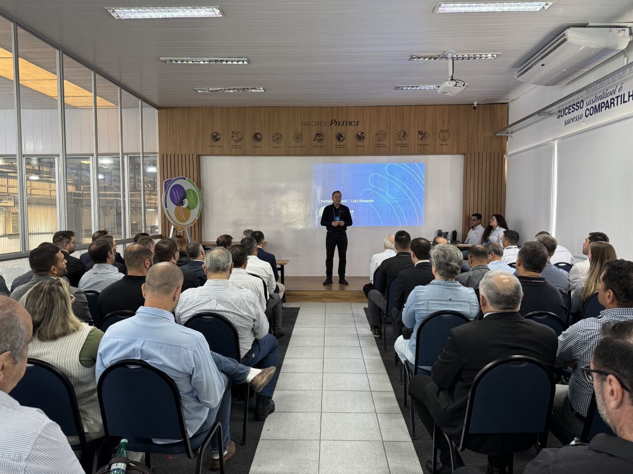 Homem fazendo uma apresentação para uma plateia em uma sala de reuniões com uma tela de projeção.