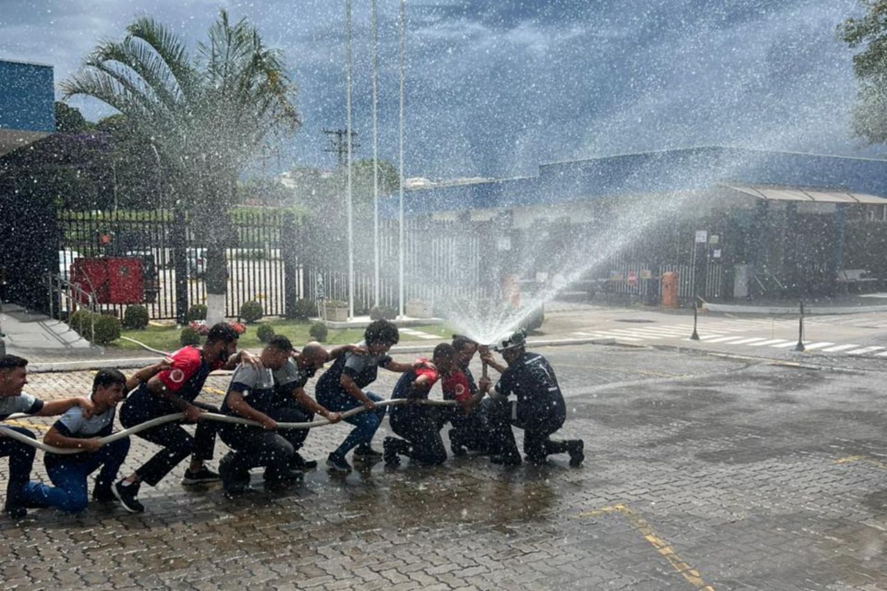 Bombeiros praticam com mangueira, lançando água. Ao ar livre, perto de prédios, alguns segurando a mangueira.