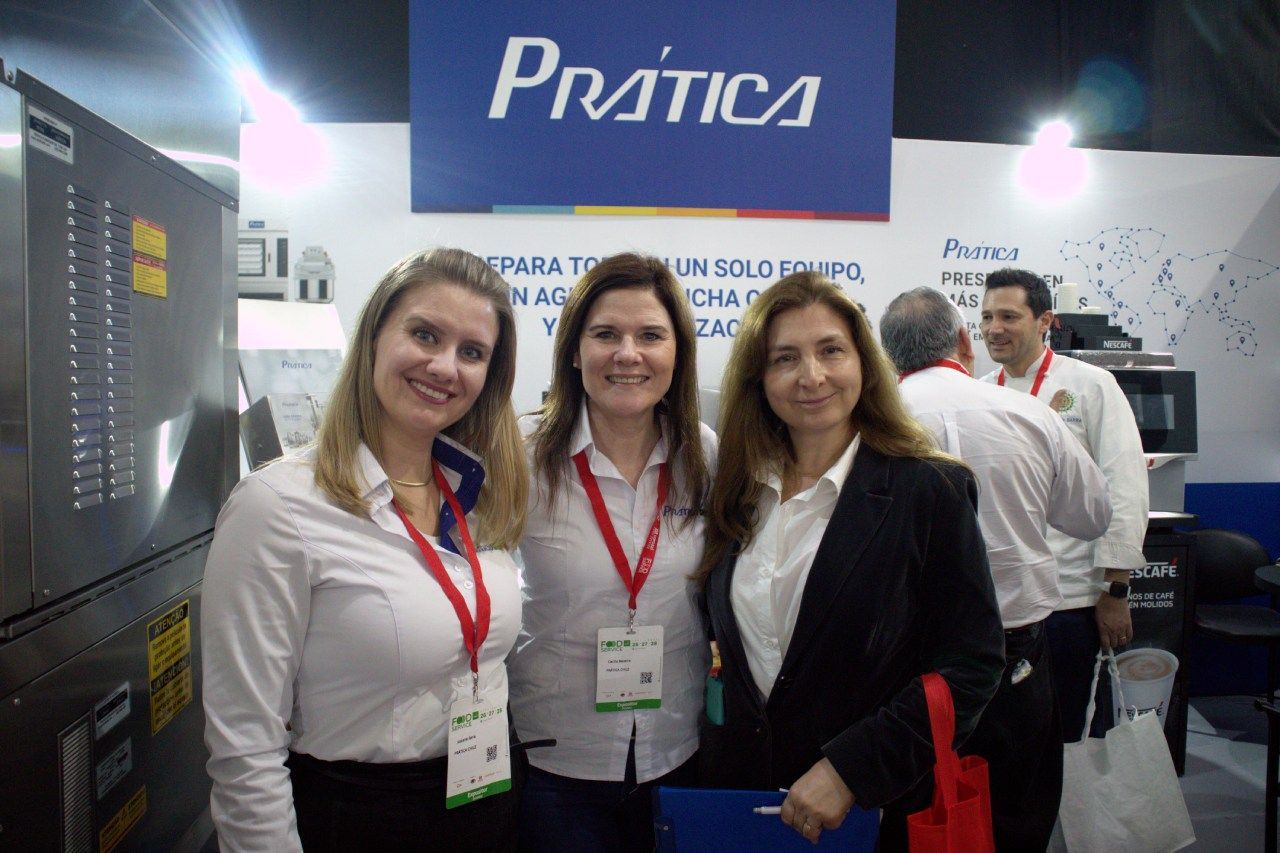 Três mulheres posam para uma foto em frente a uma placa que diz pratica