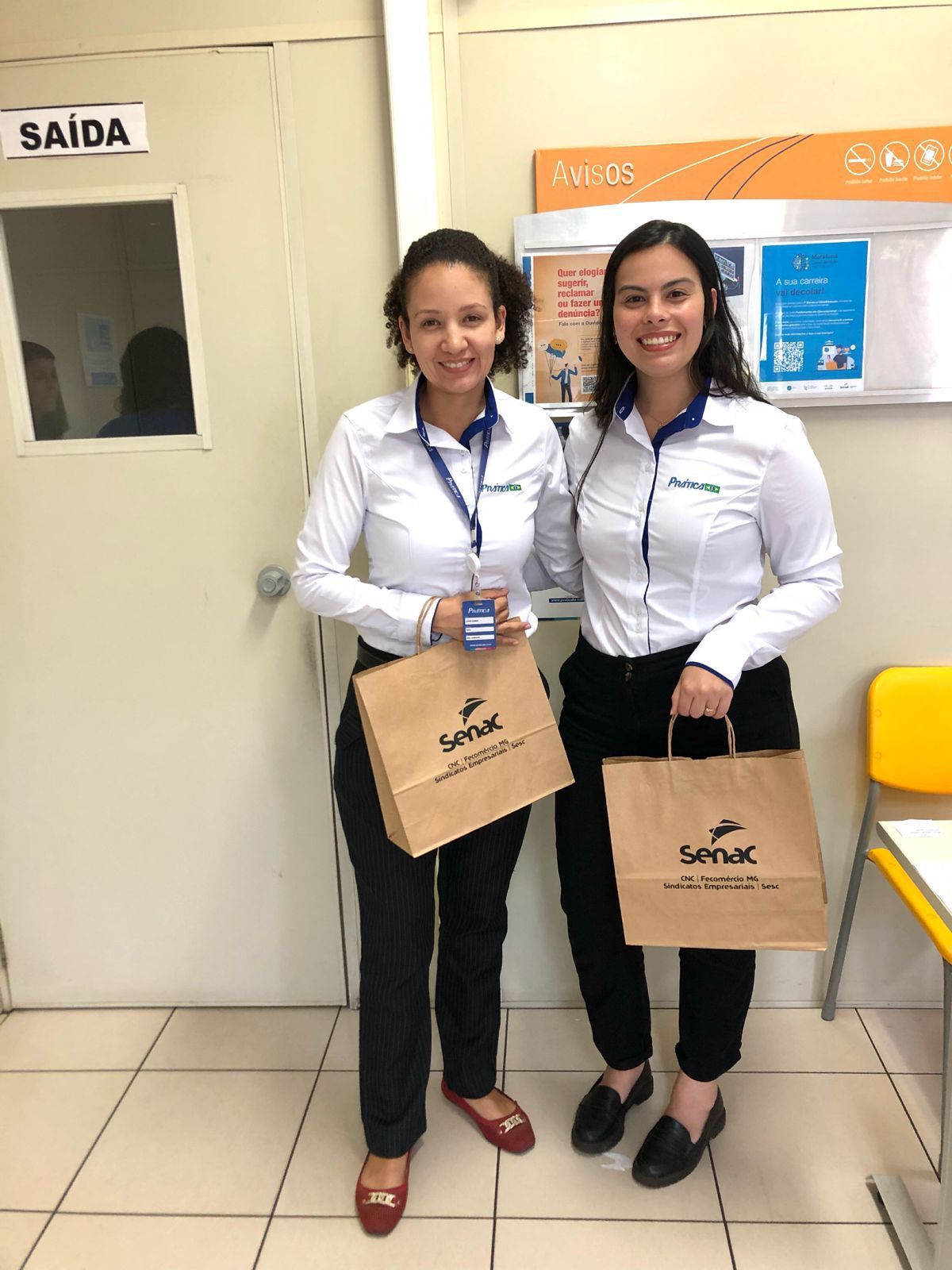 Duas mulheres estão de pé, uma ao lado da outra, em uma sala, segurando sacolas.