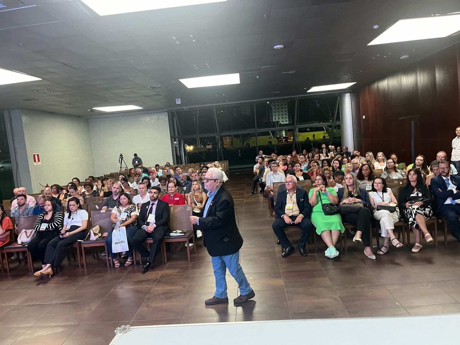 Um homem está de pé na frente de um grande grupo de pessoas sentadas em cadeiras.