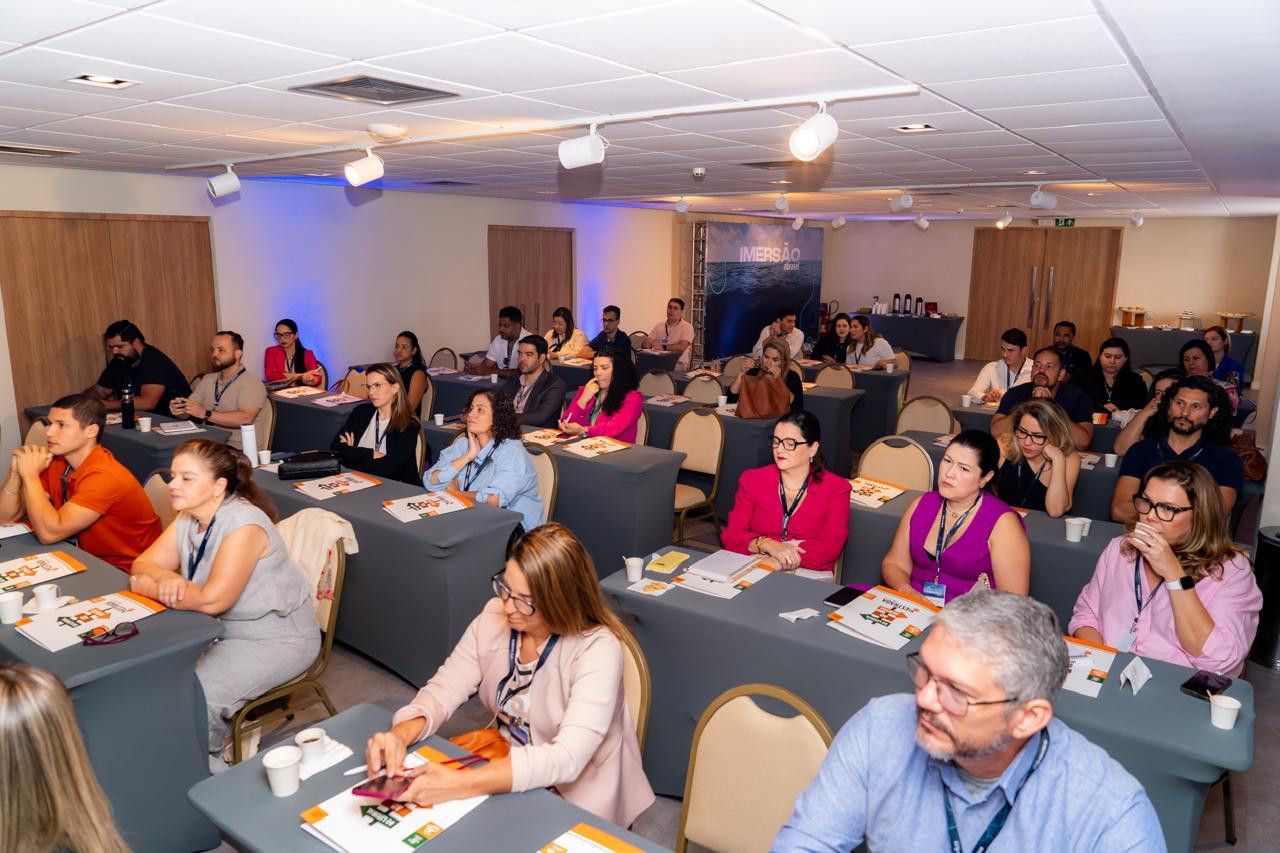 Um grande grupo de pessoas está sentado em mesas em uma sala de conferências.
