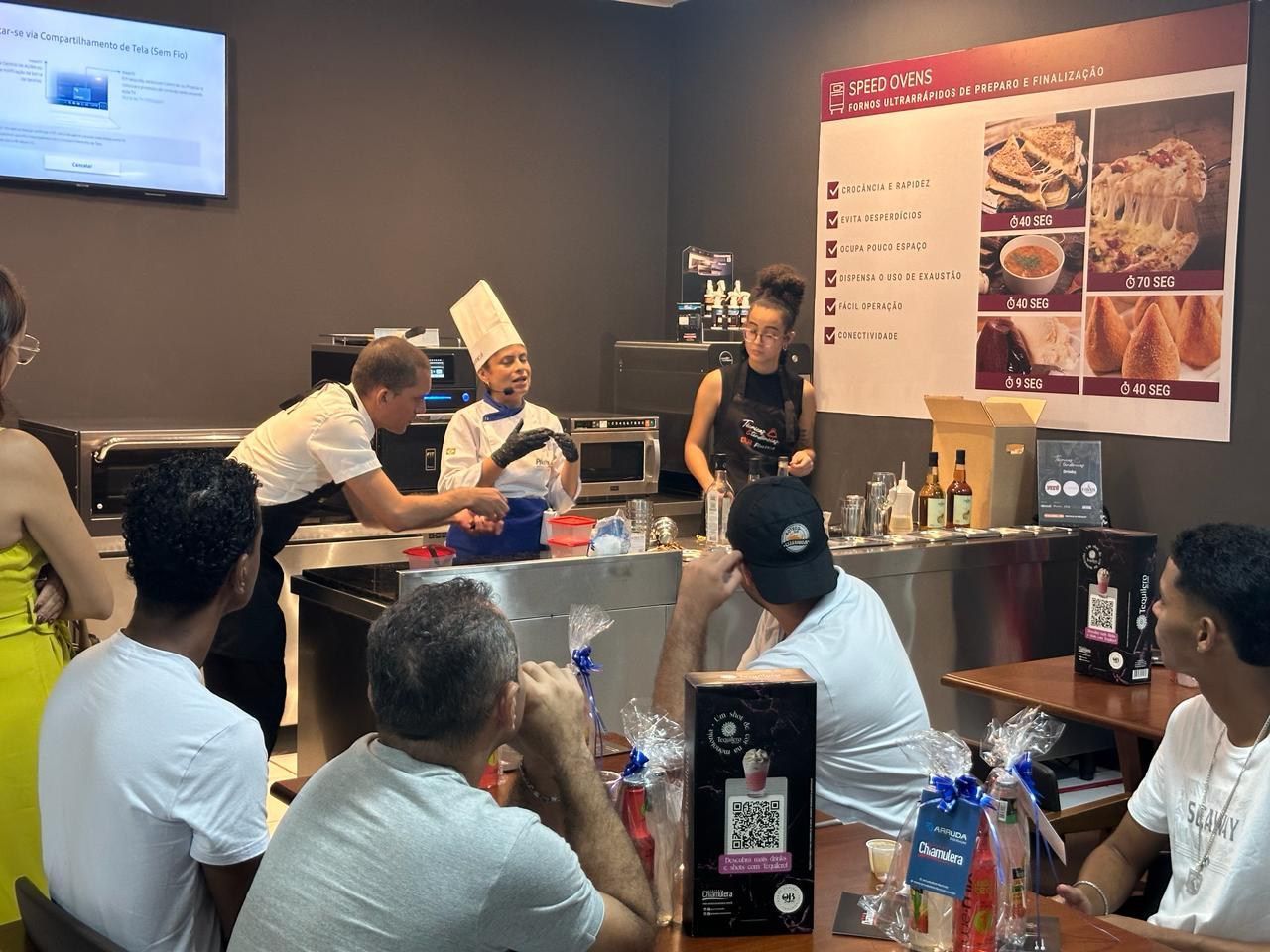 Um grupo de pessoas está sentado em mesas em uma cozinha assistindo a uma aula de culinária.