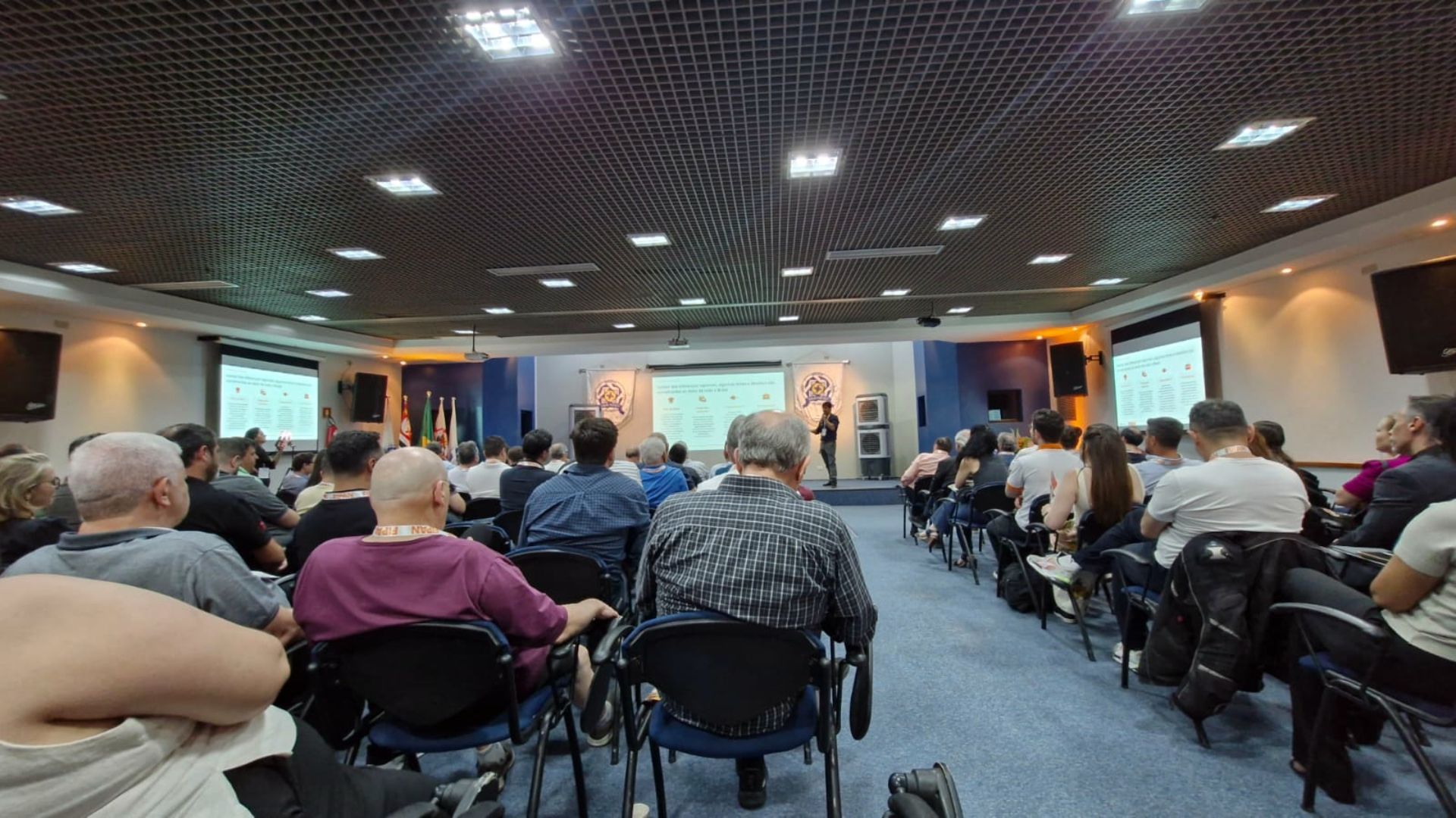 Público em uma apresentação em uma sala de conferências com paredes azuis; apresentador em pé diante de uma tela.