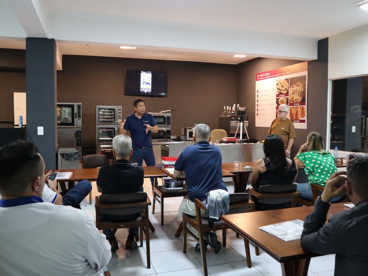 Um homem está fazendo uma apresentação para um grupo de pessoas sentadas em mesas em uma sala.