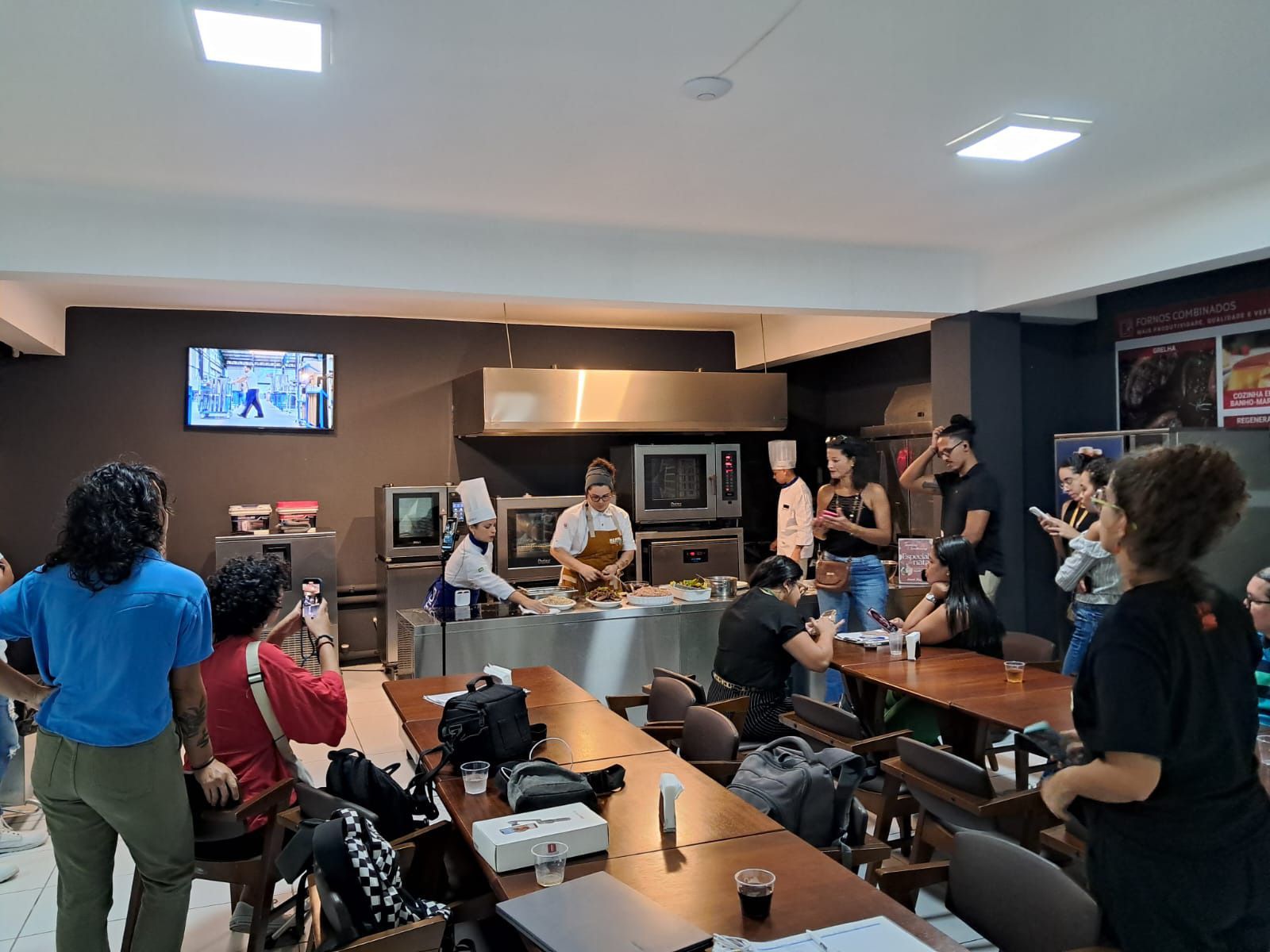 Um grupo de pessoas está sentado em mesas em uma cozinha assistindo a uma aula de culinária.