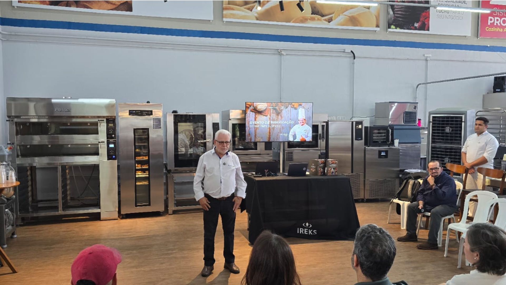 Homem faz apresentação em demonstração de equipamento de padaria; público assiste.