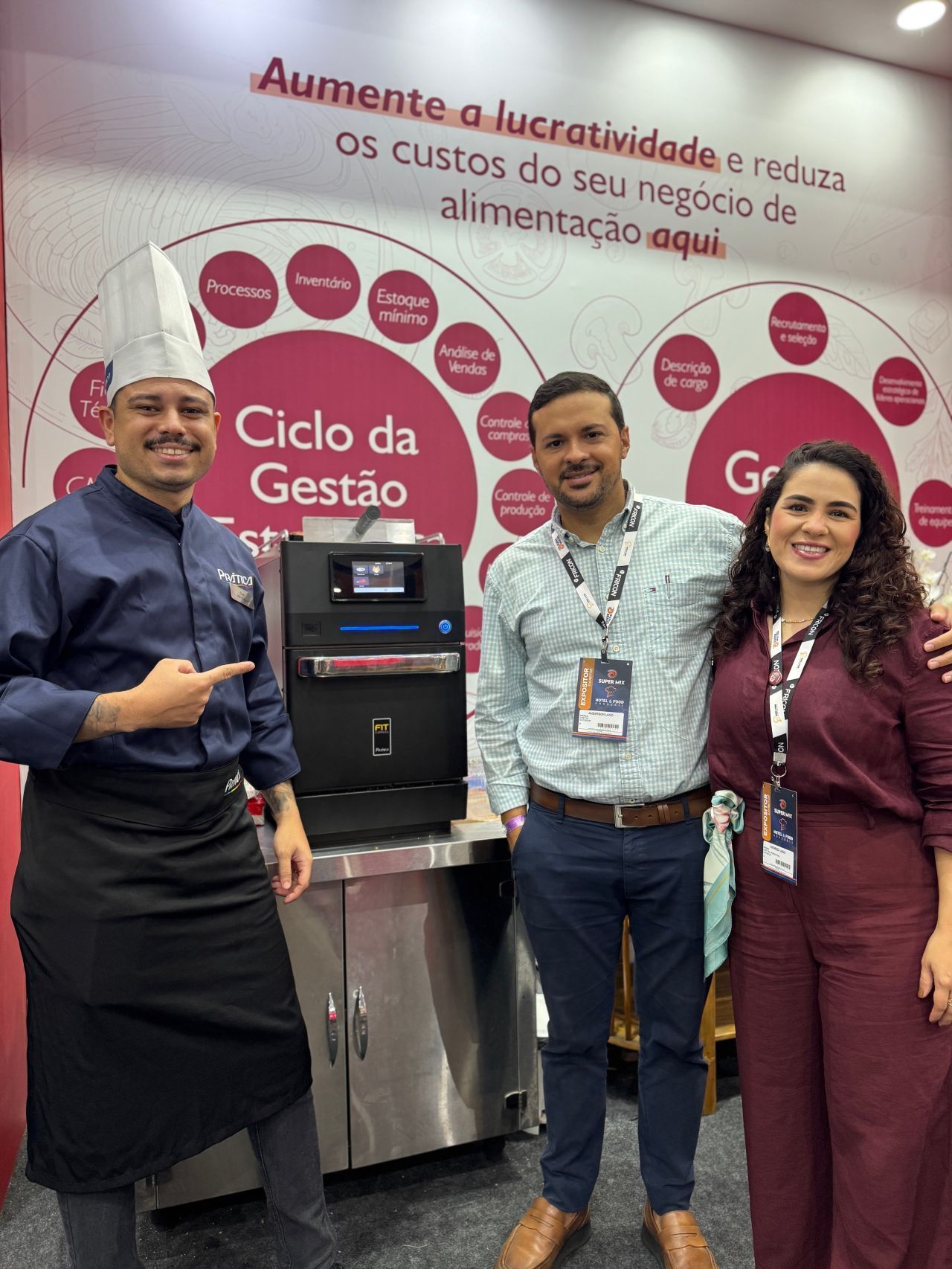 Três pessoas junto a um equipamento de serviço de alimentação em um estande de feira comercial, com um gráfico de ciclo de gestão ao fundo.