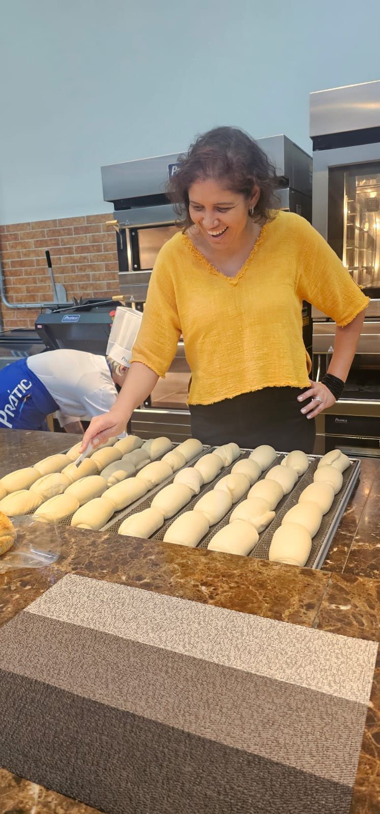 Uma mulher de camisa amarela está parada em frente a uma bandeja de massa.