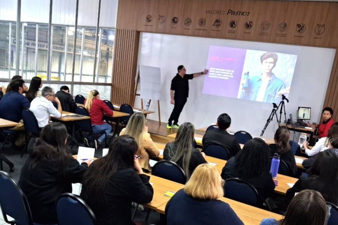 Um homem faz uma apresentação para uma turma. A apresentação mostra uma pessoa de paletó, com texto. Os alunos estão sentados, de frente para o apresentador.