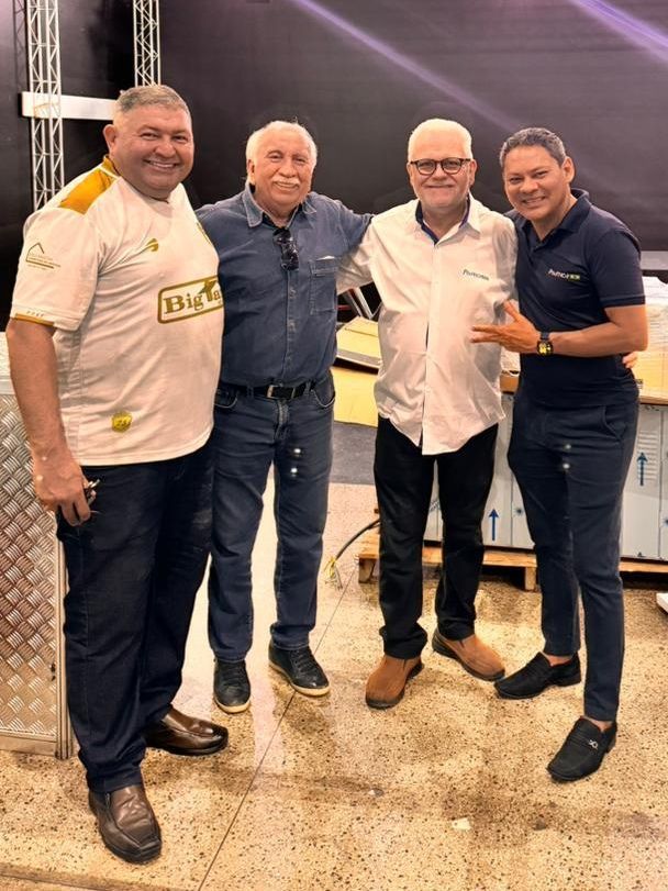 Quatro pessoas posam juntas, sorrindo. Homens em roupas casuais, um de malha, outro de jeans, em um ambiente interno.
