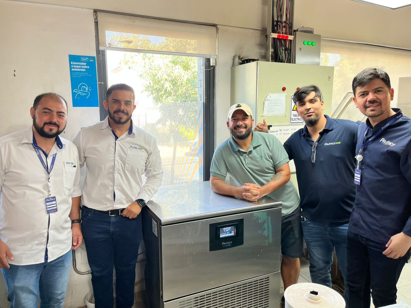 Cinco homens posam sorrindo ao lado de uma máquina de aço inoxidável em um ambiente industrial.