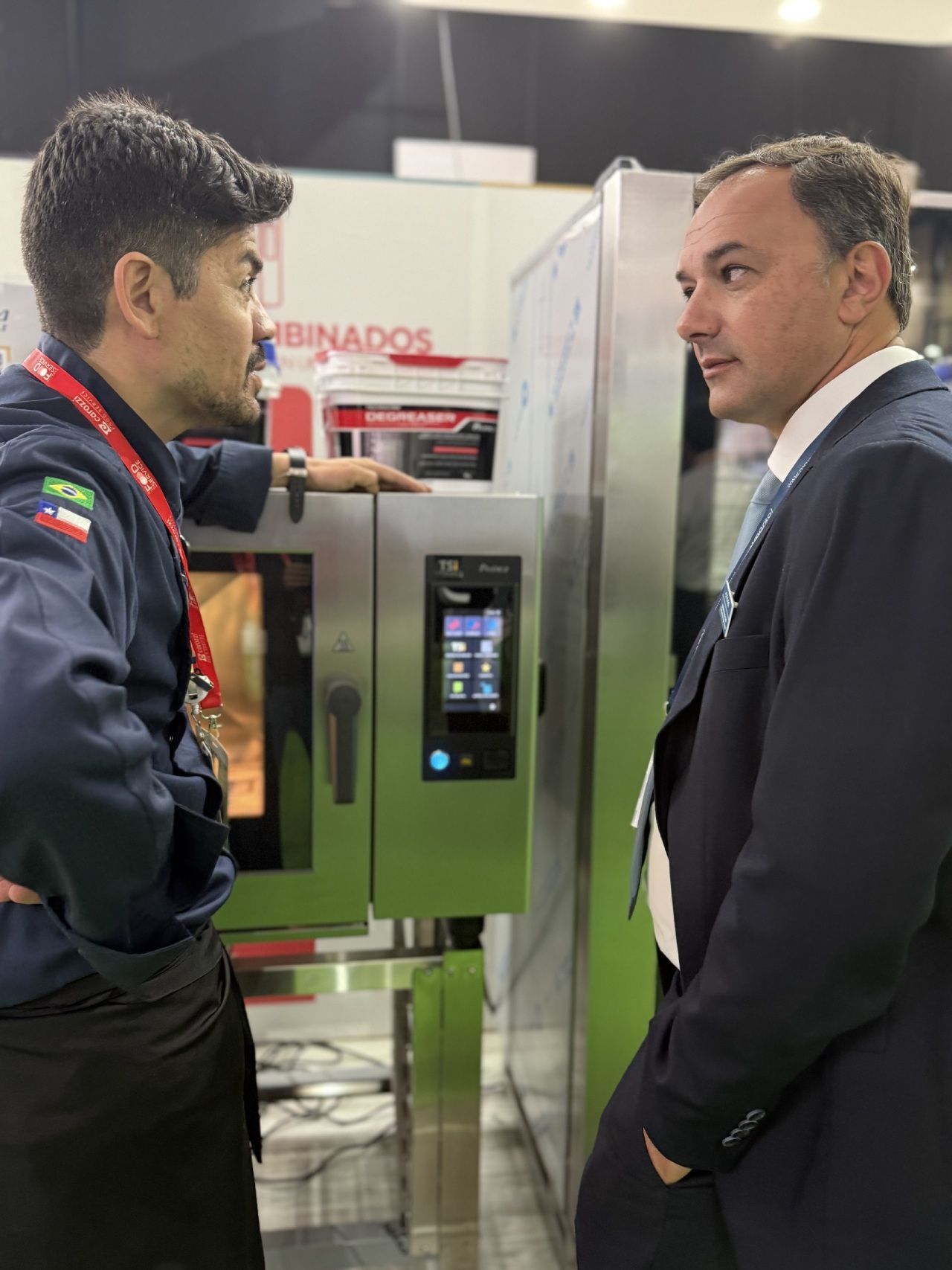 Chef de uniforme azul e homem de terno discutem sobre forno em uma feira comercial.