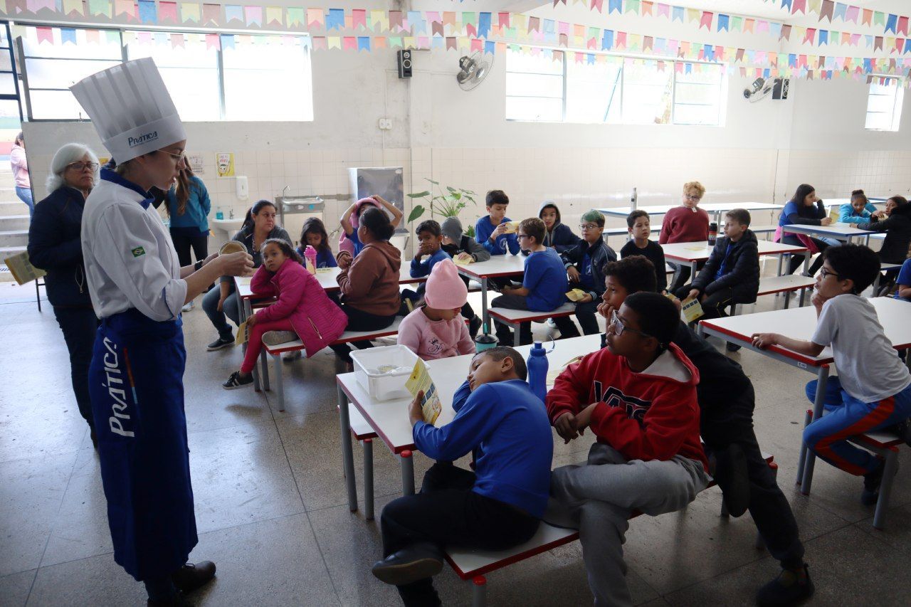 Um homem com chapéu de chef está conversando com um grupo de crianças no refeitório de uma escola