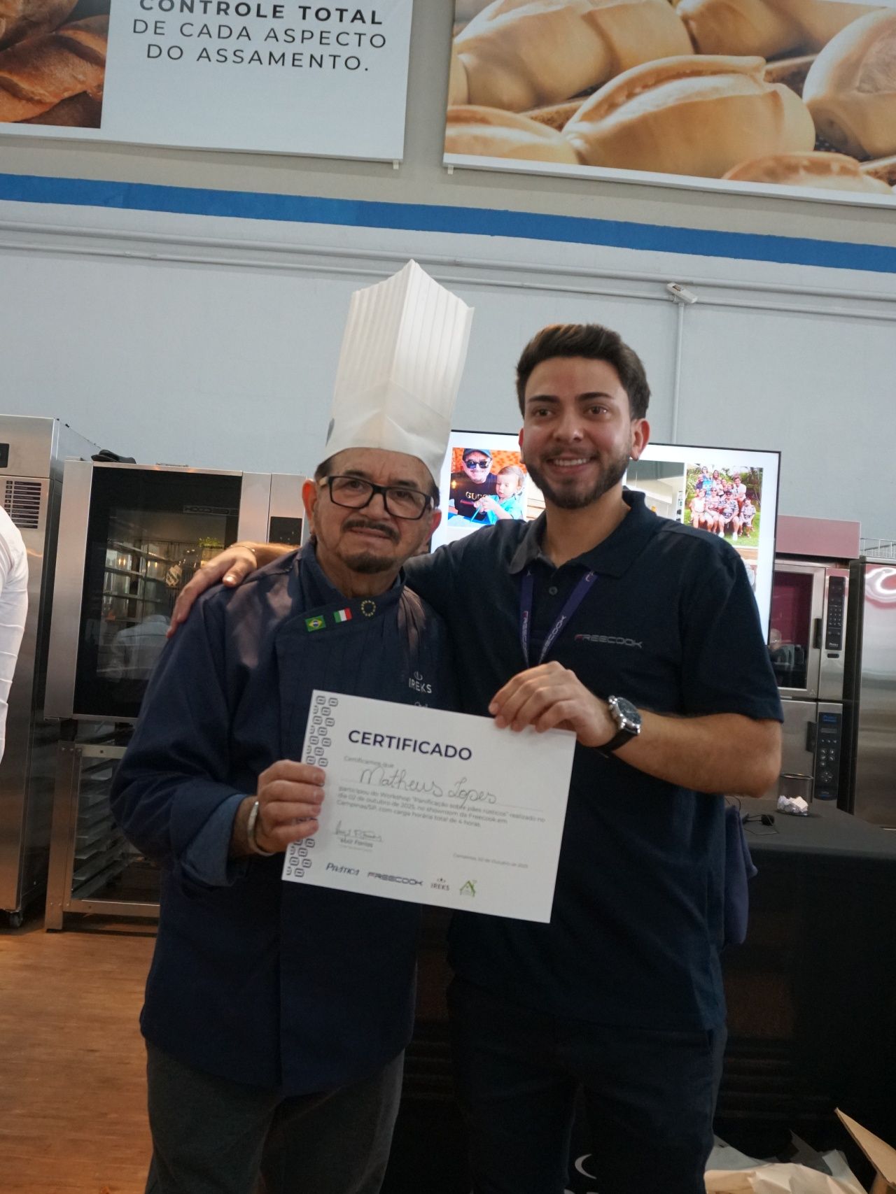 Dois homens posando com um certificado em uma padaria. Um homem mais velho usa chapéu e uniforme de chef, sorrindo. O outro homem sorri e abraça o homem mais velho.