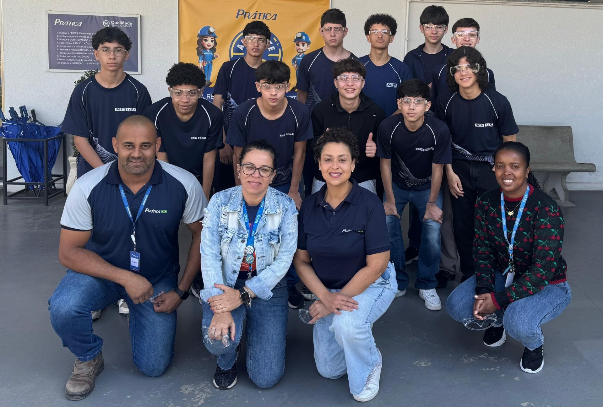 Foto de grupo: adolescentes, adultos, ao ar livre, camisas azuis, polegares para cima, fundo de prédio, dia ensolarado.