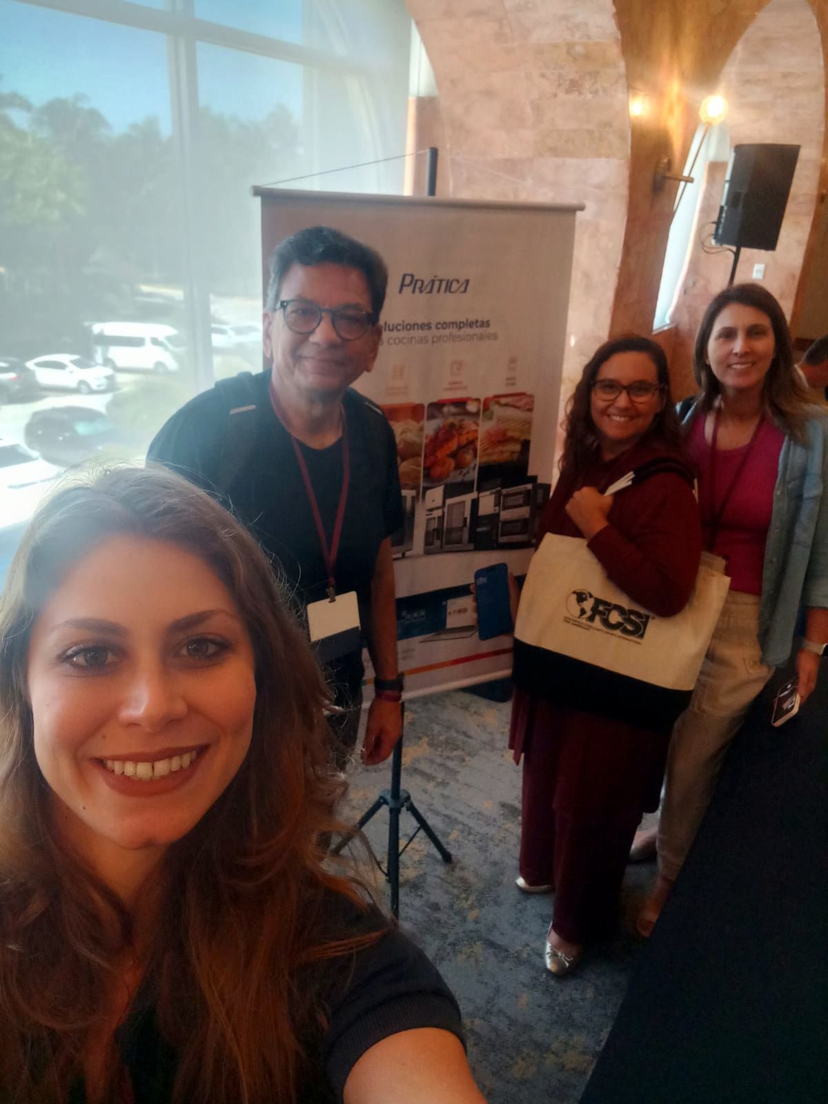 Selfie em grupo em uma conferência. Quatro pessoas posam perto de um banner de vendedor; a luz do sol entra.