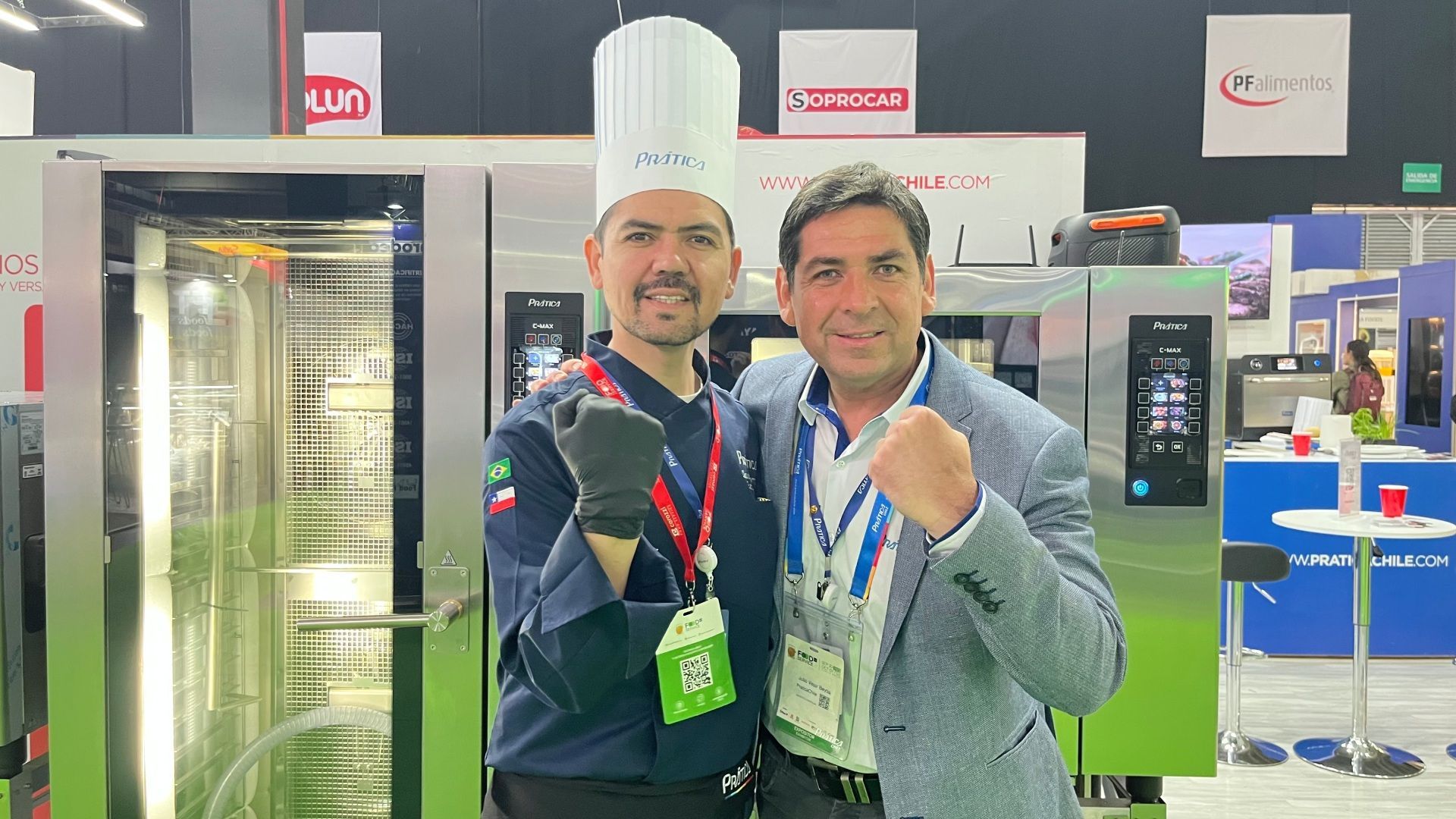 Dois homens posam em frente a fornos comerciais em uma feira. Um deles usa touca e punho cerrado.