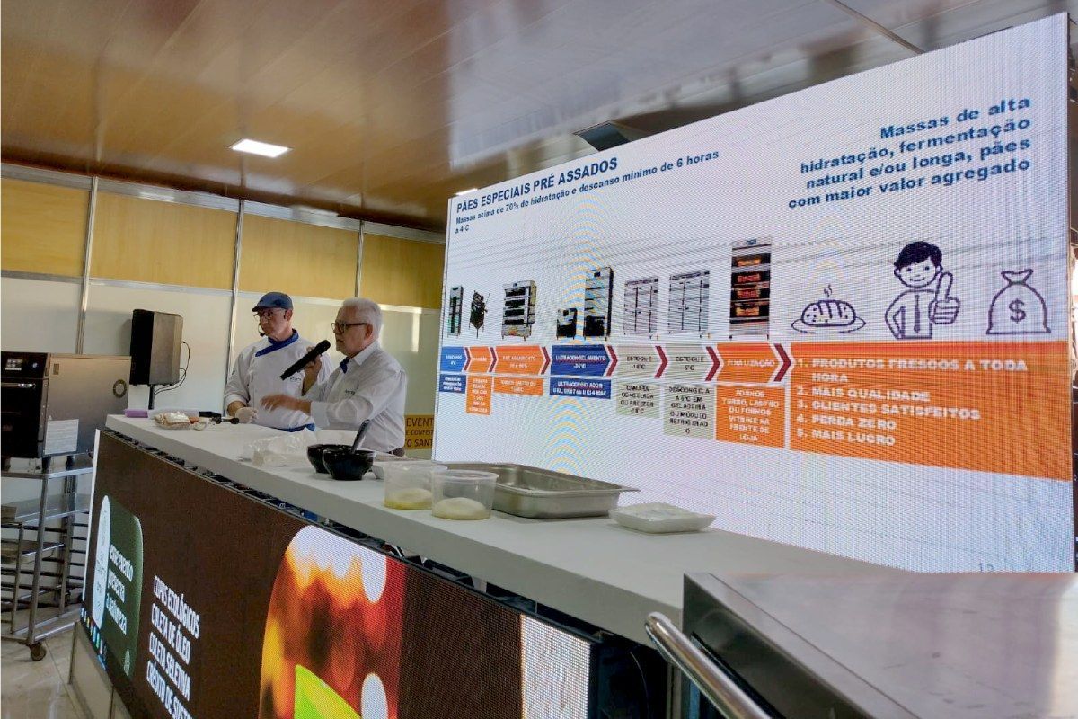 Dois homens estão de pé atrás de um balcão em frente a uma tela grande.