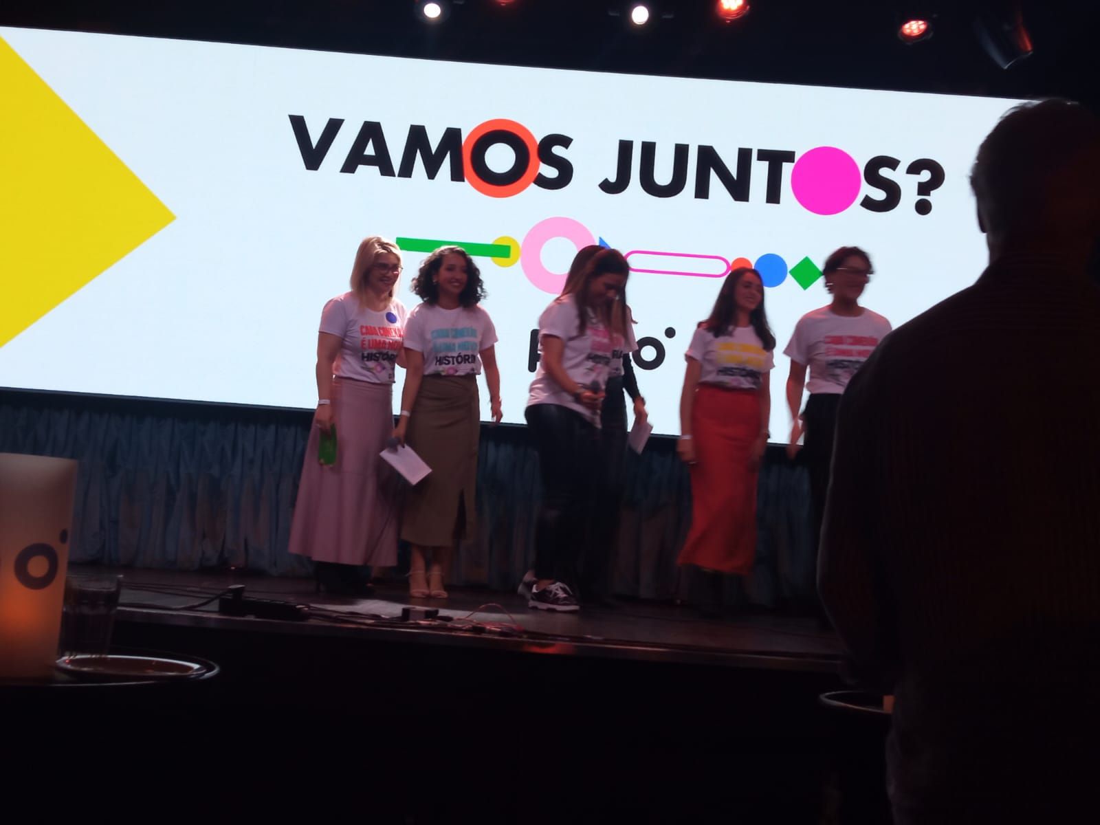 Um grupo de mulheres está em um palco em frente a uma placa que diz vamos juntos