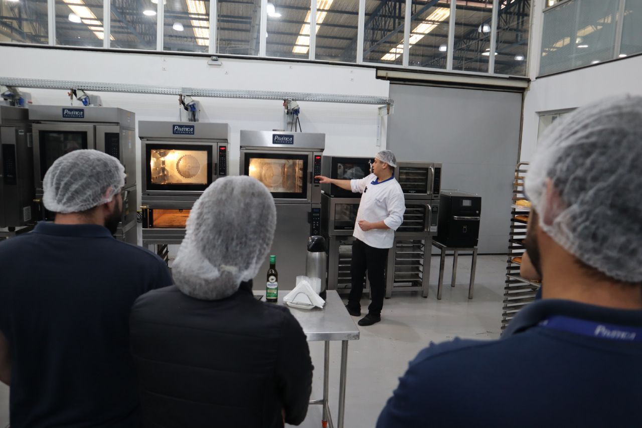 Um homem com um chapéu branco está conversando com um grupo de pessoas em uma cozinha.