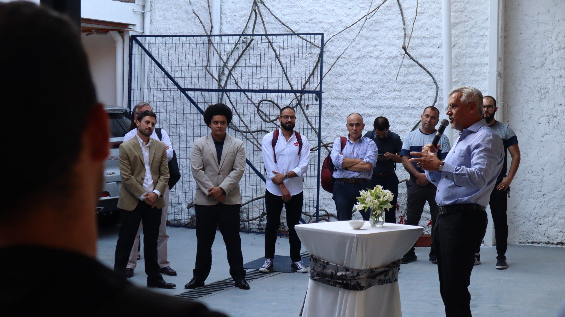 Um grupo de homens está de pé ao redor de uma mesa enquanto um homem fala em um microfone.