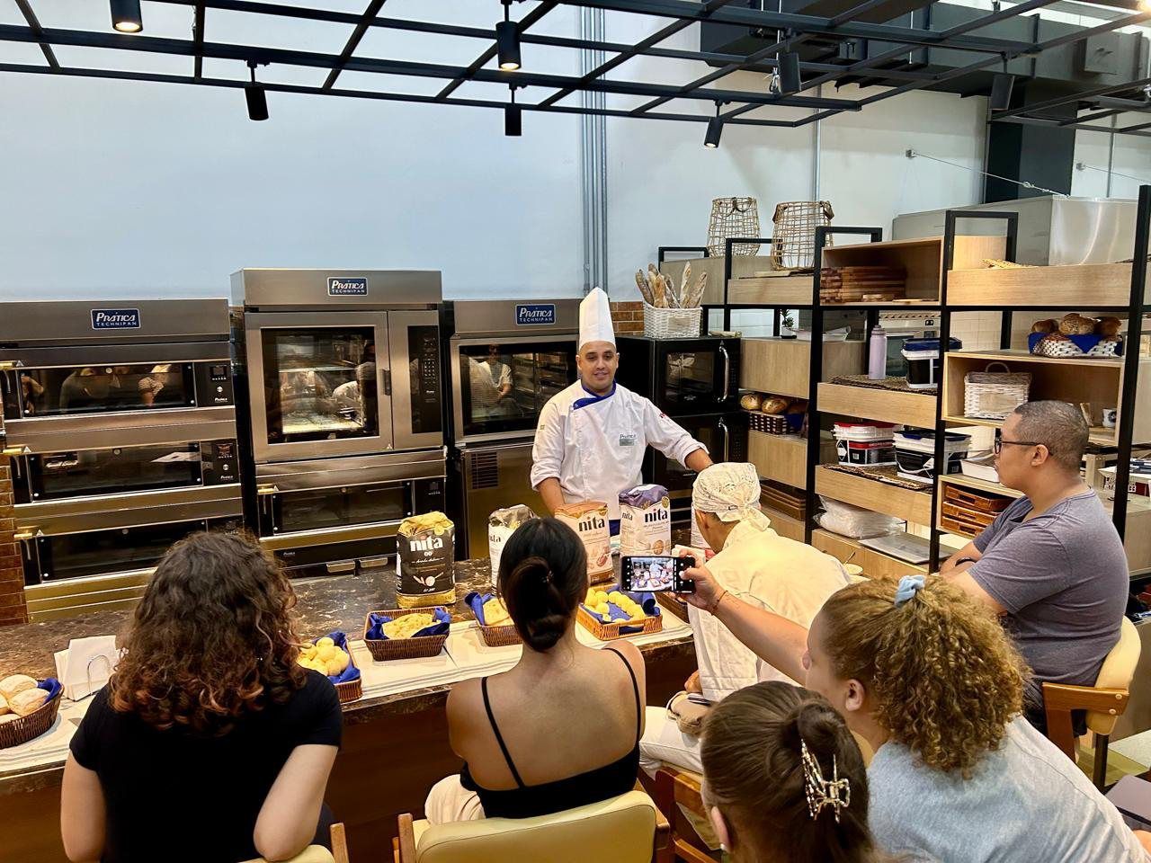 Um grupo de pessoas está sentado à mesa em frente a um chef em uma cozinha.
