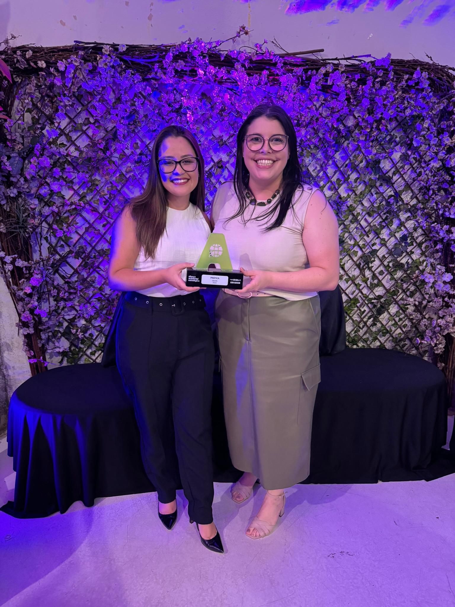 Duas mulheres estão de pé, uma ao lado da outra, segurando um troféu.